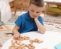 A boy arranging wooden letters on a table while resting his chin on his hand