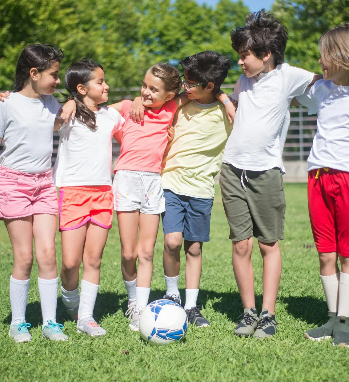 A group of children in sports kit standing together outdoors with a football at their feet